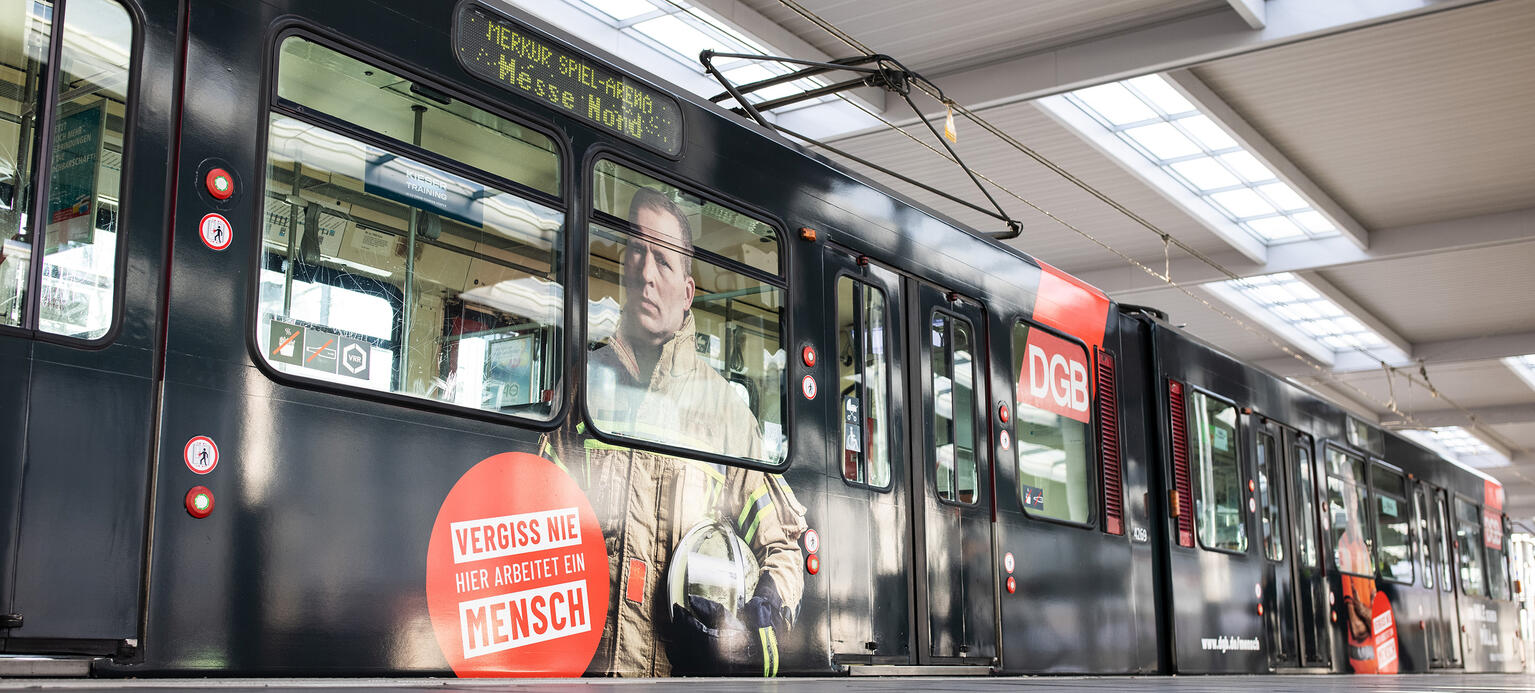 Eine Stadtbahn mit der Aufschrift „Vergiss nie hier arbeitet ein Mensch“ des Deutschen Gewerkschaftsbunds (DGB) steht an einer Haltestelle.